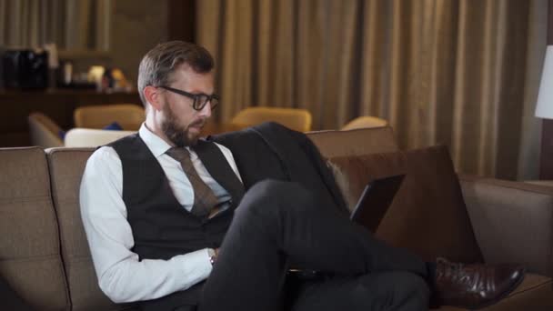 Nuori Liikemies Laseissa Työskentelee Kannettavassa Tietokoneessa Mies Puvussa Istuu Sohvalla — kuvapankkivideo