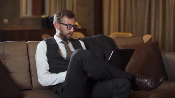 Młody Biznesmen Okularach Pracujący Laptopie Mężczyzna Garniturze Siedzący Kanapie Pokoju — Wideo stockowe