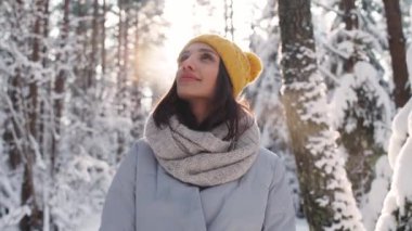 Kış ormanındaki güzel bir genç kadının portresi, neşeli bir kız doğadaki bir kış yürüyüşü sırasında poz veriyor ve kameraya bakıyor, 4k yavaş çekim..