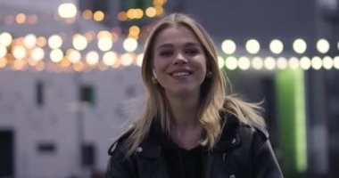 Gülümseyen ve poz veren güzel bir kızın portresi, modern yerleşim alanı, şehirde akşam, arka planda bulanık ışıklar, neşeli ve pozitif duygular..