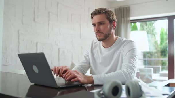 ハンドヘルド ワイヤレスヘッドフォンに焦点を当てた男は 机の上に自宅に座って リモートワーク上のラップトップ トレーダーを使用して動作します — ストック動画