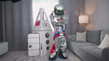 Astronot kostümü giymiş Asyalı bir genç uzay gemisinin mukavva modelinin yanında duruyor ve kaskını takıyor, kameraya bakıyor ve dalgalara bakıyor, bir çocuk oturma odasında astronotu oynuyor..