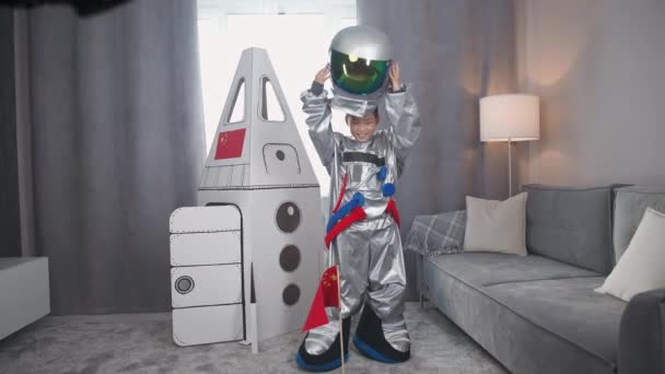 身穿航天服的亚洲少年男孩站在一艘纸板箱模型的宇宙飞船旁边 戴上头盔 看着相机和海浪 男孩在客厅里扮演航天员 — 图库视频影像