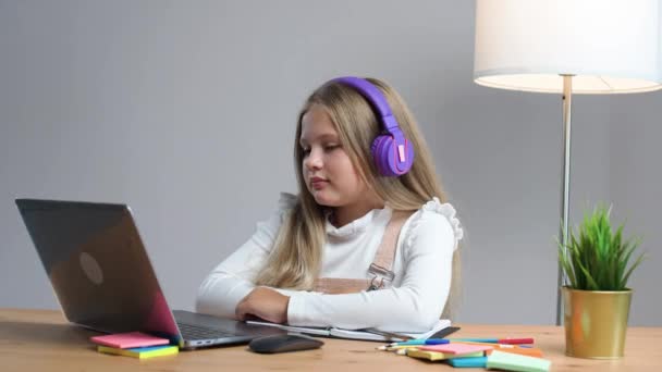 遠隔学習 家庭のテーブルに座っている紫色のヘッドフォンの女子高生は オンラインで自己隔離 ホームスクーリングで勉強するためにラップトップを使用します — ストック動画