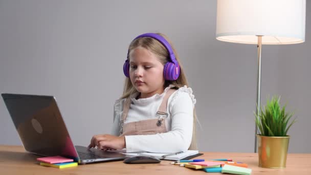 オンラインでホームスクーリング 家庭のテーブルに座っている紫色のヘッドフォンの女子高生は 自己隔離で勉強するためにラップトップを使用します — ストック動画