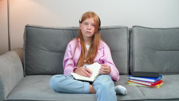 通过视频 远距离学习 坐在客厅沙发上的红头发女孩在家学习 然后在笔记本上写字 在家自学 — 图库视频影像