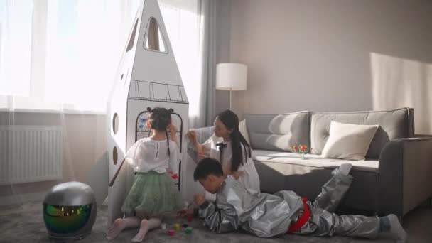 子供を持つアジアの女性が自宅のリビングルームで遊ぶ 彼女の母親と妹と一緒に床に横たわっている宇宙飛行士の衣装の男の子 子供たちは段ボールモデルに母親のペイントと一緒に — ストック動画