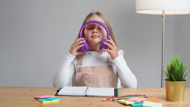 オンラインでホームスクーリング 家庭でテーブルに座って自己隔離で勉強する紫色のヘッドフォンの女子 — ストック動画