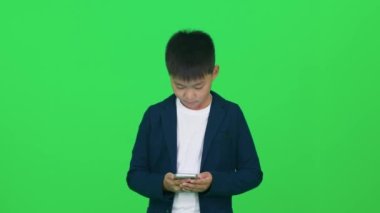 Odaklanmış Asyalı okul çocuğu yeşil arka planda duruyor ve akıllı telefon kullanıyor, bir kromatonda şablon kullanıyor..