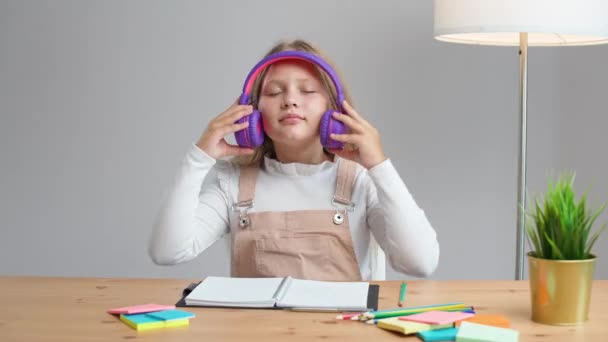 オンラインでホームスクーリング 家庭でテーブルに座って自己隔離で勉強する紫色のヘッドフォンの女子は ヘッドフォンを置きます — ストック動画