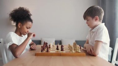 Evdeki çocuklar, odaklanmış koyu tenli bir kız ve beyaz bir oğlan oturma odasında oturmuş satranç oynuyorlar, eğlence, düşünce süreci..