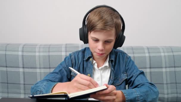 ホームスクーリング ビデオ通話でオンライン遠隔学習 リビングルームの職場に座って 自宅で自己隔離で勉強するためにウェブカメラを使用して ヘッドフォンの少年はノートブックに書きます — ストック動画
