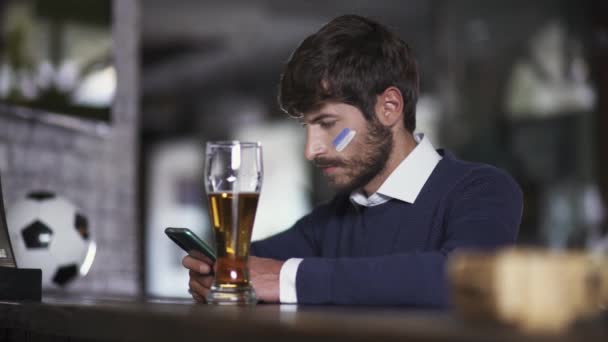 サッカーファンはサッカーの試合中にバーに座っていて スマートフォンを使用して 男は携帯電話を使ってテキストを読んでいます — ストック動画