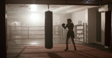 Kadın boksör yumruklarını eğitir, kum torbasını döver, boks salonunda antrenman yapar, arka ışıklandırma, boks yapar..