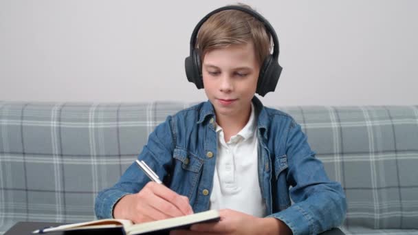ホームスクーリング ビデオ通話でオンライン遠隔学習 リビングルームの職場に座って 自宅で自己隔離で勉強するためにウェブカメラを使用して ヘッドフォンの少年はノートブックに書きます — ストック動画