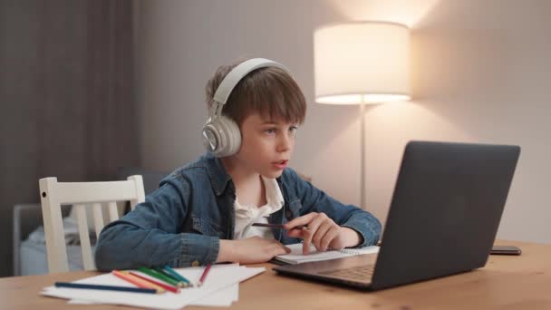 ビデオ通話による距離学習 リビングルームに座ってラップトップを使用して自宅で自己隔離で勉強 ヘッドフォンの少年はオンラインで通信します — ストック動画