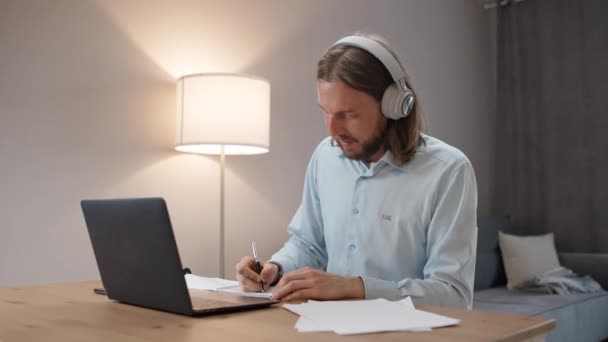 リモートワークのフリーランサー ビデオ会議でのオンライン会議でヘッドフォンの男 リビングルームでの職場 リモートジョブで人 男はノートブックに書く — ストック動画