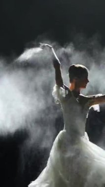Dramatik dans, dikey video, beyaz etekli zarif bir kadın, balerin dans ediyor ve siyah arka planda koreografik öğeler sergiliyor, magnezya kullanıyor, karanlık bir sahnede sis etkisi yaratıyor..