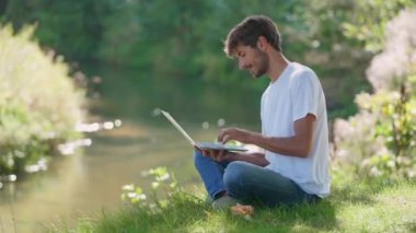 Nehir kenarında otururken dizüstü bilgisayar başında çalışan, yazı yazan ve doğada uzaktan kumandalı iş yapan bir adam tatildeyken çalışır..