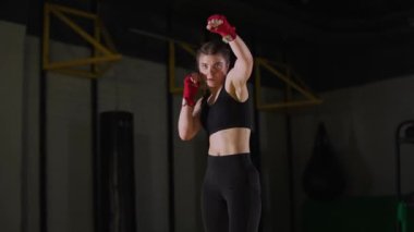 Sportif kadın dövüşçü savunmasını eğitiyor, boks salonunda antrenman yapıyor, genç kadın kameraya bakıyor..