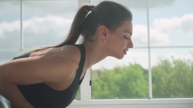 Işıkla dolu beyaz bir odada yoga yapan genç bir kadın büyük pencerenin yanında kaslarını gerer..