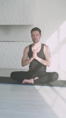 Genç adam meditasyon yapıyor ve yoga yapıyor, sakin bir ruh hali, ışıkla dolu beyaz bir odada dinleniyor, adam yerde oturuyor, sosyal ağlar için dikey video..