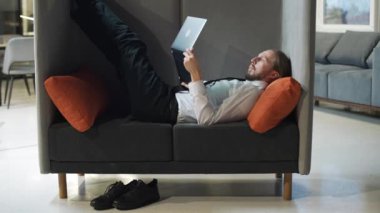 Ofis müdürü büyük bir sandalyede uzanıyor ve dizüstü bilgisayarda çalışıyor, ofis işi açık alanda, bir adam dizüstü bilgisayarına mesaj yazıyor, rutin bir iş..