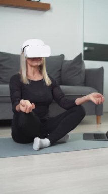 Sanal gerçeklik gözlüklü neşeli yaşlı kadın oturma odasında meditasyon yapıyor, modern teknoloji ve artırılmış gerçeklik, bir kadın kendini tecrit, farkındalık, dikey video.