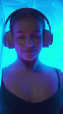 Kulaklıklı neşeli kadın duyusal yoksunluk odasında yatıyor, mavi neon ışığı, spa, portre, dikey video.
