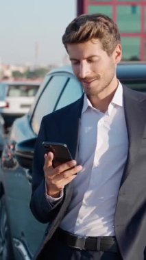 Takım elbiseli genç işadamı yeni arabanın yanında duruyor ve akıllı telefon kullanıyor. Neşeli adam yeni bir arabaya biniyor. Kiralık araba, dikey video..