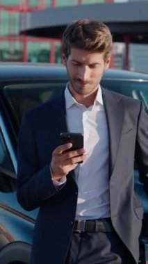 Takım elbiseli bir işadamı yeni arabanın yanında duruyor ve akıllı telefon kullanıyor. Neşeli adam yeni bir arabaya biniyor. Kiralık araba, dikey video..