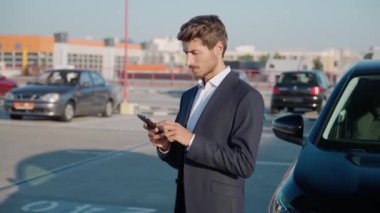 Takım elbiseli genç işadamı yeni arabanın yanında duruyor ve akıllı telefon kullanıyor, neşeli bir adam, kiralık araba kullanıyor..
