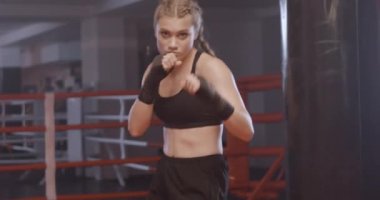Kadın boksör yumruklarını eğitiyor, boks salonunda antrenman yapıyor, kız bir dizi yumruk eğitiyor ve kameraya bakıyor..