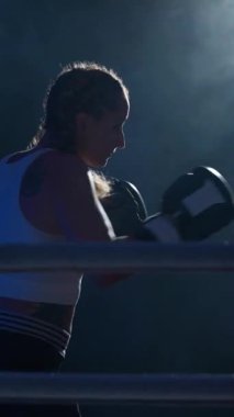 Yavaş çekim dikey video, kadın dövüşçü kum torbasına vuruyor, boks salonunda antrenman yapıyor, yumruklarını eğitiyor..