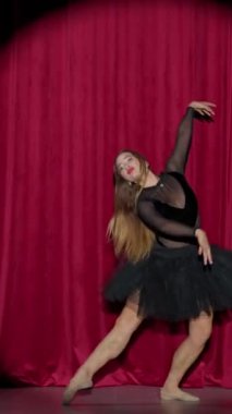 Dikey video, dramatik performans, siyah etekli güzel balerin kırmızı arka planda, sahne ışığı altında dramatik dans gösterisi..