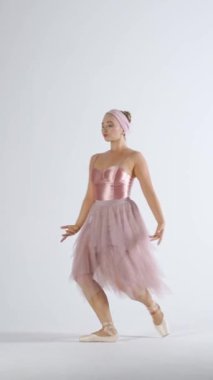 Pembe etekli bir balerin dans eder ve beyaz arka planda, dikey bir videoda dramatik koreografik öğeler sergiler..