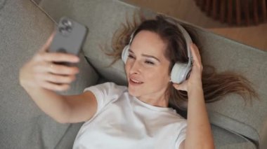 Neşeli bir kadın kanepede uzanıyor ve akıllı bir telefonla video selfie çekiyor, kulaklıkla müzik dinliyor..
