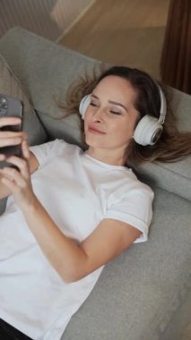 Dikey video, kanepede uzanmış neşeli bir kadın akıllı bir telefonla video selfie çekiyor, kulaklıkla müzik dinliyor..