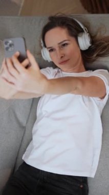 Neşeli bir kadın kanepede uzanıyor ve akıllı bir telefonla video selfie çekiyor, kulaklıkla müzik dinliyor, dikey videolar çekiyor..
