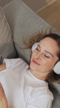Elde tutulan, neşeli bir kadın kanepeye uzanıyor ve akıllı bir telefonla video selfie çekiyor, kulaklıkla müzik dinliyor, dikey videolar dinliyor..