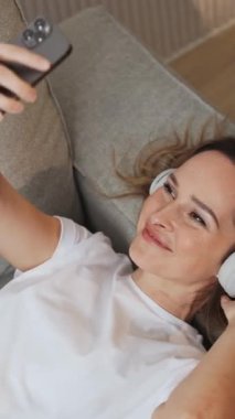 Dikey video, kanepede uzanmış neşeli bir kadın akıllı bir telefonla video selfie çekiyor, kulaklıkla müzik dinliyor..