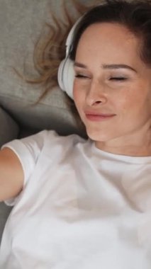 Dikey video, kanepede uzanmış neşeli bir kadın akıllı bir telefonla video selfie çekiyor, kulaklıkla müzik dinliyor..