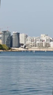 Malta, Valletta - 23 Mart 2025: Modern şehir manzarası ve ufuktaki küçük yatlar, mimari ve yat limanı, dikey video manzarası, deniz manzarası.