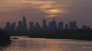 Varşova, Polonya - 18 Nisan 2025: Akşam vakti hızlandırılmış şehir manzarası, ufukta gökdelenlerin siluetleri, nehir ve günbatımı gökyüzü manzarası, hızlı hareket, gece vakti iş merkezi.