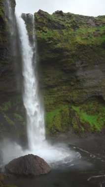 İzlanda, bir şelalenin içinde, İzlanda doğasının dikey videosu, dinginlik, su akışı manzarası, yeşil kayalık arazi arka planı.
