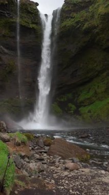 İzlanda, bir şelalenin içinde, İzlanda doğasının dikey videosu, dinginlik, su akışı manzarası, yeşil kayalık arazi arka planı.