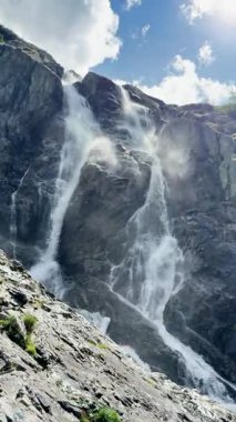 Dikey video, dağ manzarası, devasa şelale manzarası, dingin şelale manzarası, kayalık arazi arka planı, eriyen buzullar ve akan su..