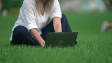 Ağır çekimde, yeşil alanda oturan ve laptopla çalışan kimliği belirsiz bir kadın. Şehir parkında dizüstü bilgisayar kullanan yetişkin bir öğrenci..