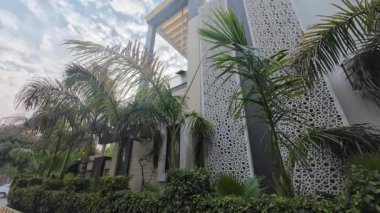 Bulutlu bir gökyüzünün altındaki palmiye ağaçlarıyla çevrili modern bir evin yan görünüşü çağdaş dış tasarımını sergiliyor..