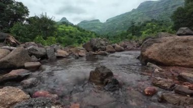 Ormandaki kayaların üzerinden akan sakin bir akarsu manzarası, doğanın rahatlatıcı seslerini ve canlı yaprakları sergiliyor.
